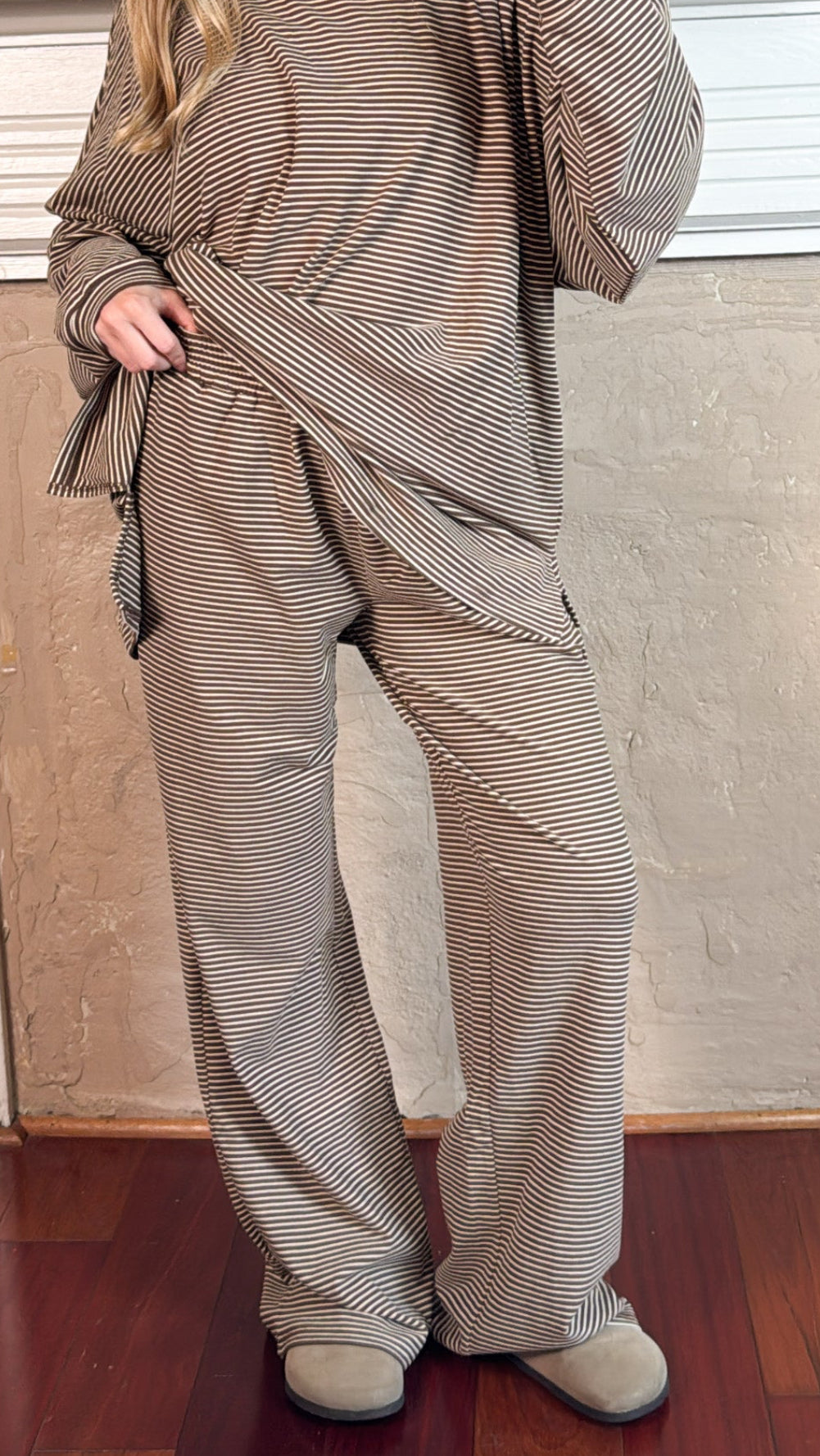 A woman takes a mirror selfie wearing a matching two-piece lounge set with a fine horizontal stripe pattern in beige and brown. The set includes a long-sleeve top and wide-leg pants, styled with beige slippers in front of a fireplace.